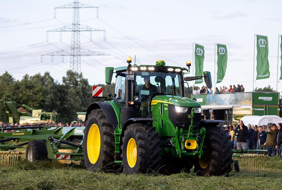 John Deere Traktor mit KRONE Schwader auf Grünland. Menschen, die der Maschine bei der Arbeit zusehen im Hintergrund.